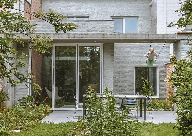 Projectbeeld Woning J&H Kortrijk: aanbouw met lichte beigegele baksteen tegen witte stadswoning, mensen op terras met hond achter glazen schuifdeur - warm en huiselijk.